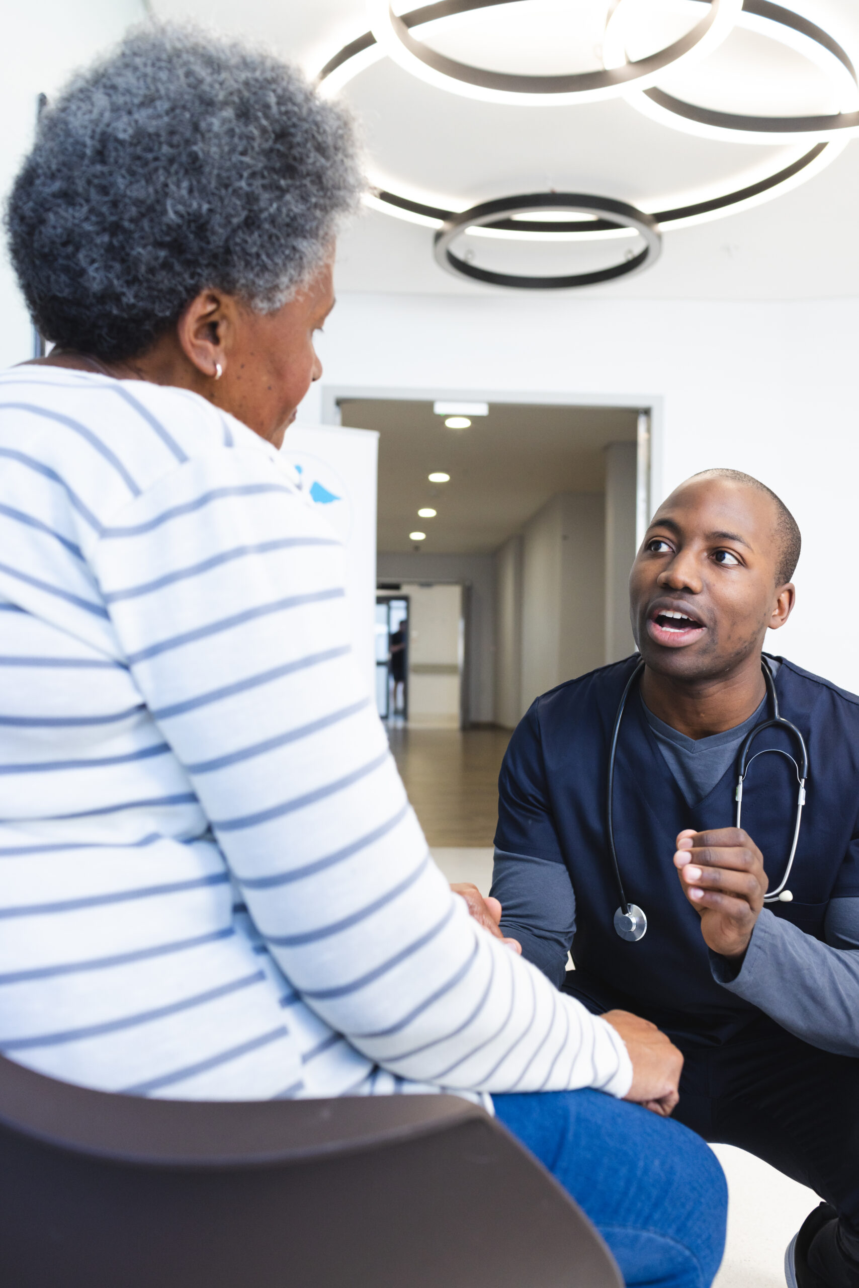 African american male doctor talking with senior woman in hospital waiting room. Medicine, healthcare and medical services, unaltered.