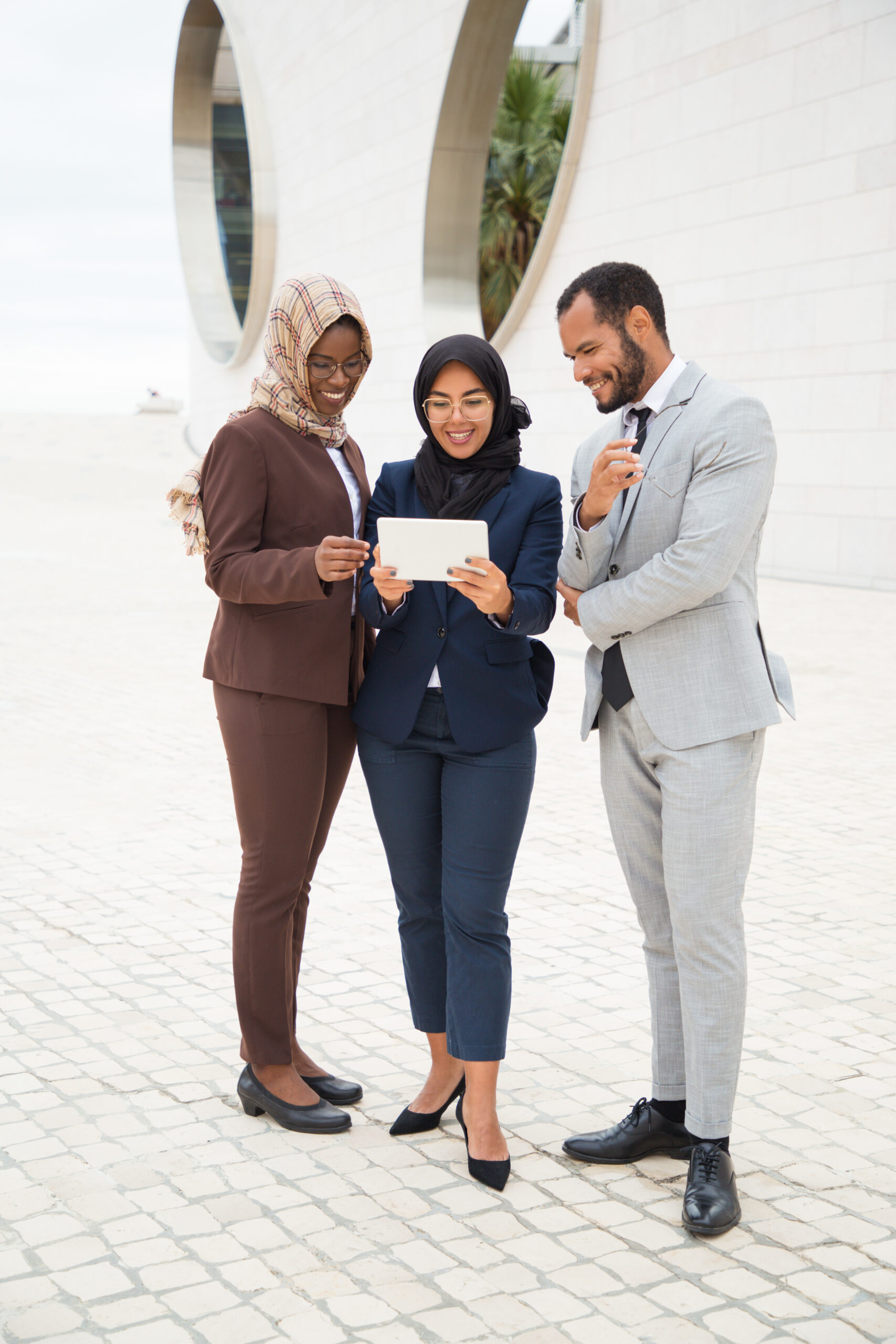 Joyful diverse business colleagues watching funny videos on tablet outside. Businessman and Muslim businesswomen standing outdoors and using tablet together. Media content concept